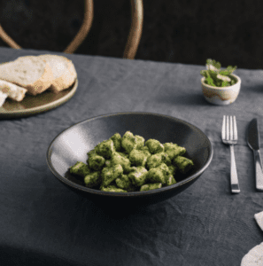 A black bowl filled with basil pesto gnocchi sits atop a table laid with a charcoal tablecloth, cutlery and a plate of sliced bread.