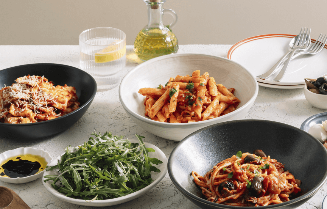 A table laid with bowls filled with different types of pasta: spaghetti primavera, penne amatriciana, beef ravioli bolognese and rocket salad.