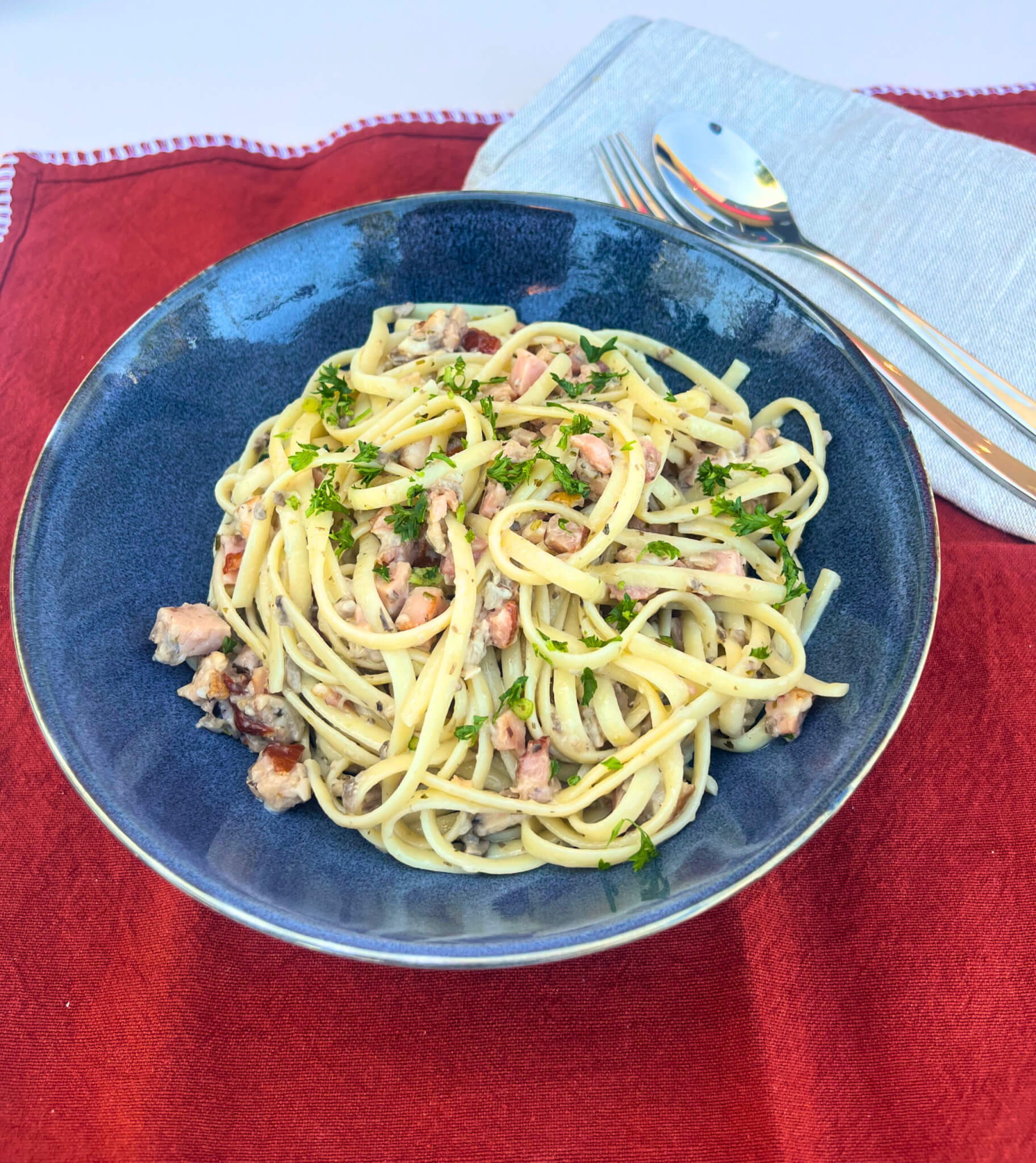 Creamy Bacon and Mushroom Linguine plated up in a blue bowl on a red tablecloth with a napkin and cutlery set.