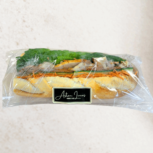 White baguette roll filled with BBQ-seasoned chicken, shredded carrot, cos lettuce, tomato, cucumber and mayonnaise and packaged in clear cellophane packaging with a black label. The baguette is at the center of the image, positioned in front of a white/grey background.