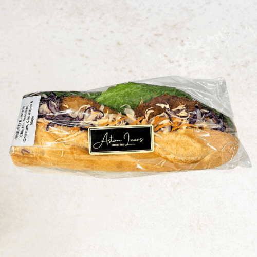 A baguette roll filled with chicken schnitzel, coleslaw, cos lettuce and mayonnaise. The baguette is packaged in clear packaging with a black logo sticker and sat atop a cream/beige background.