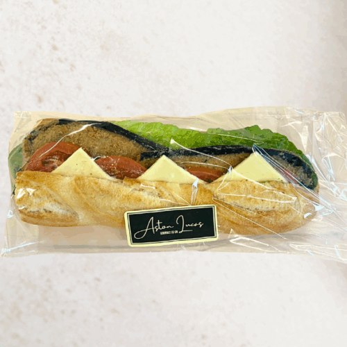 A white baguette roll filled with crumbed eggplant, tomato, cheese and cos lettuce and packaged in clear cellophane with a black label. The baguette is at the center of the image and is positioned in front of a white/grey background.