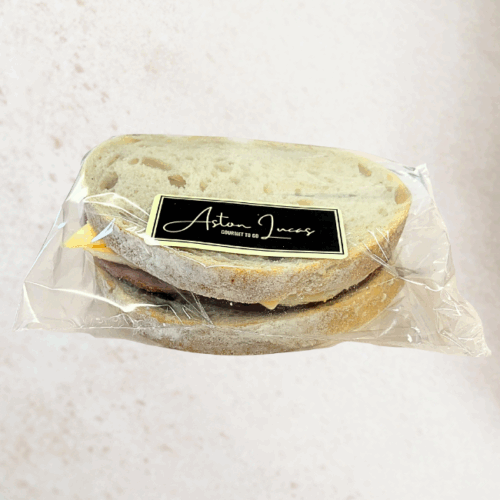 A sourdough sandwich filled with egg, bacon and cheese and packaged in clear cellophane with a black label. The sandwich is positioned at the center of the image in front of a white/grey background.