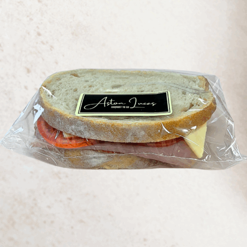 A sourdough sandwich filled with ham, tomato and mozzarella and packaged in clear cellophane with a black label. The sandwich is positioned in the center of the image in front of a white/grey background.