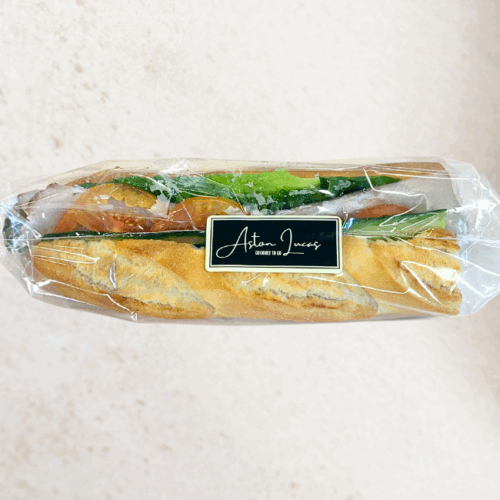 A white baguette roll filled with champagne ham, cos lettuce, tomato, cucumber and dijon mustard and packaged in clear cellophane with a black label. The baguette is at the center of the image and is positioned in front of a white/grey background.