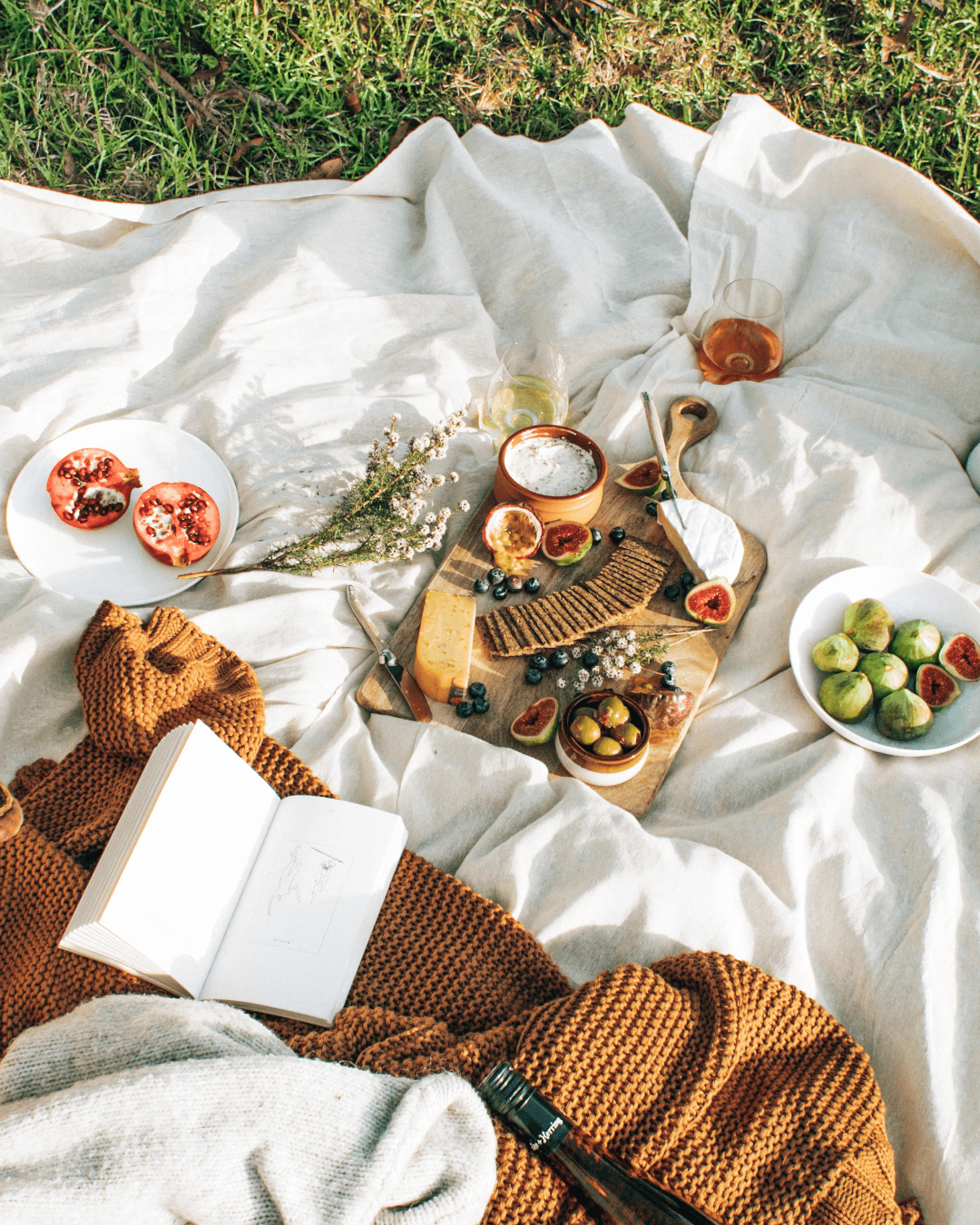 A white picnic rug on the grass is laid with a cheese platter, fruit, a book, blanket and a bottle of wine.